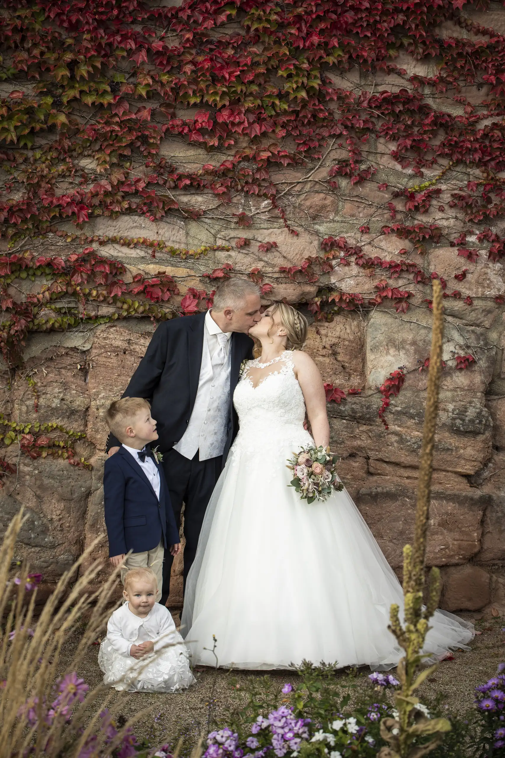 Küssendes Brautpaar mit zwei Kindern vor rotem Weinlaub im Barockgarten Heilbad Heiligenstadt.