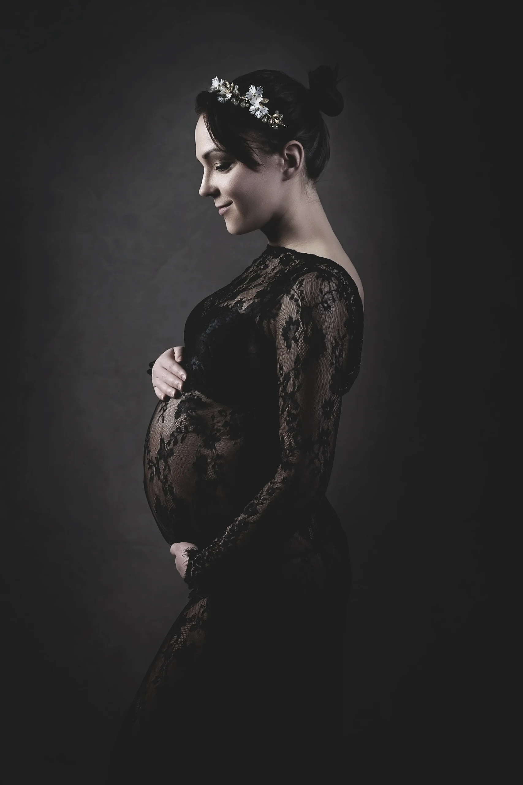 Schwangere Frau im schwarzen Spitzenkleid mit Babybauch in Seitenansicht.