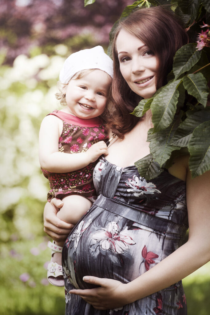Schwangere Frau mit einem Kind auf dem Arm bei einem Babybauchshooting im Garten.