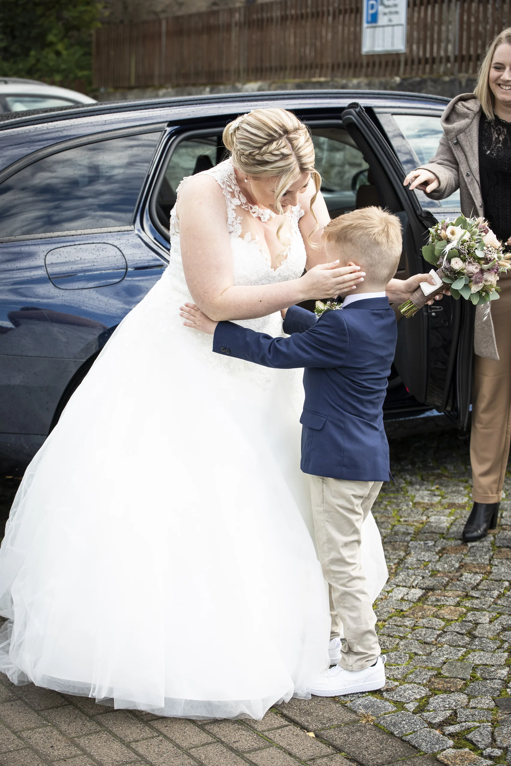 Braut umarmt einen kleinen Jungen neben dem Hochzeitsauto an der Pfarrkirche St. Gerhard in Heilbad Heiligenstadt.