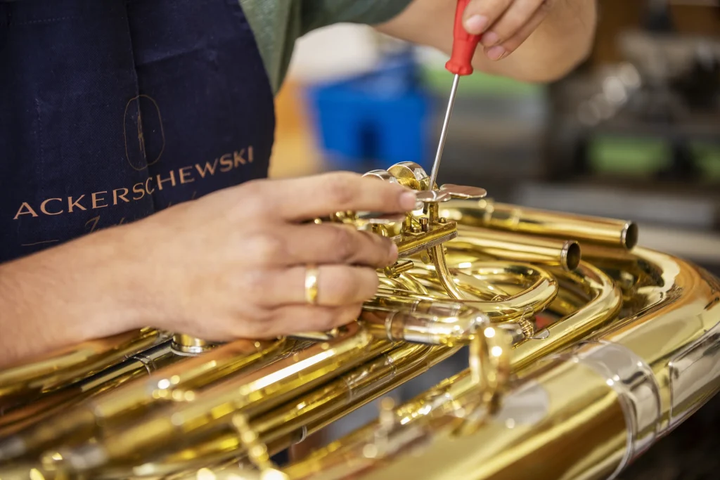Nahaufnahme von Händen, die mit einem roten Schraubendreher an der Ventilmechanik einer goldfarbenen Tuba arbeiten.