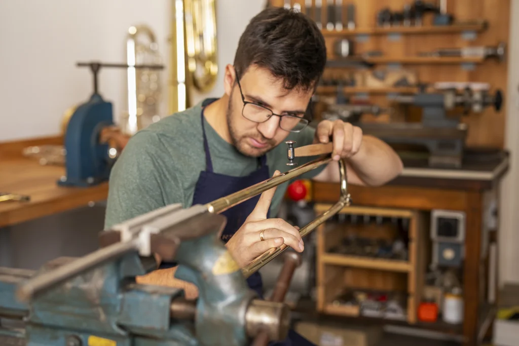 Ein Instrumentenbauer mit Brille bearbeitet ein langes Metallrohr am Schraubstock seiner Werkbank.