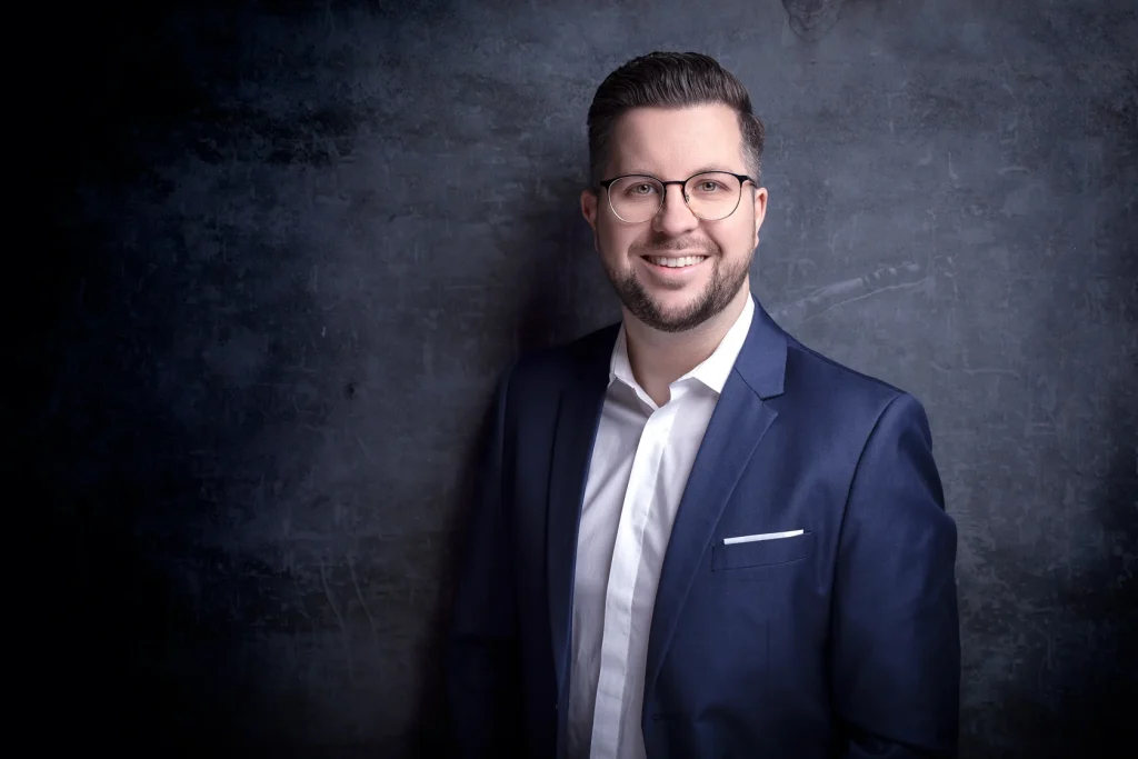 Businessporträt eines lächelnden Mannes mit Brille, dunkelblauem Sakko und weißem Hemd vor dunklem Hintergrund.