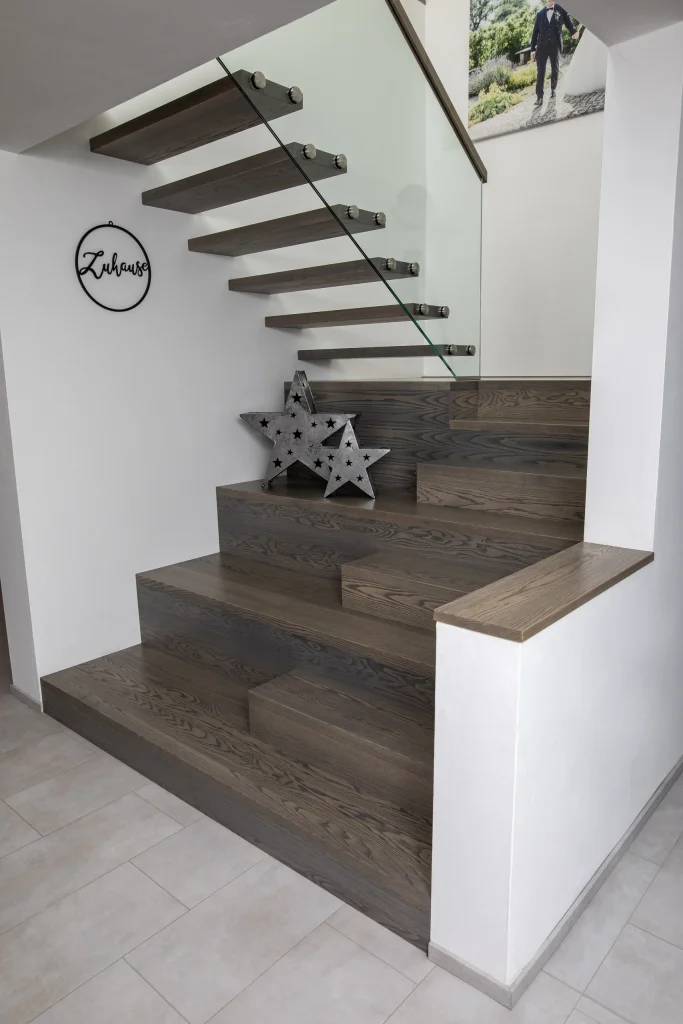 Moderne Holztreppe mit breiten unteren Stufen, frei auskragenden oberen Stufen und einem Glasgeländer.