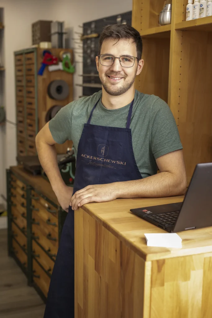 Lächelnder Handwerker mit Brille und blauer Schürze an einem Holztresen mit Laptop in seiner Werkstatt.