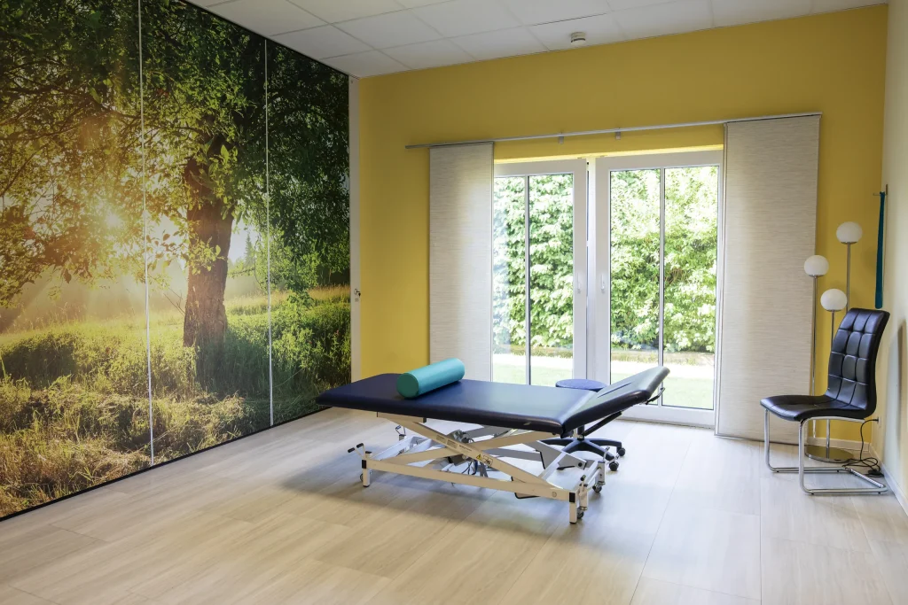 Heller Behandlungsraum mit Therapieliege, gelber Wand und großflächiger Fototapete einer sonnigen Waldlandschaft.