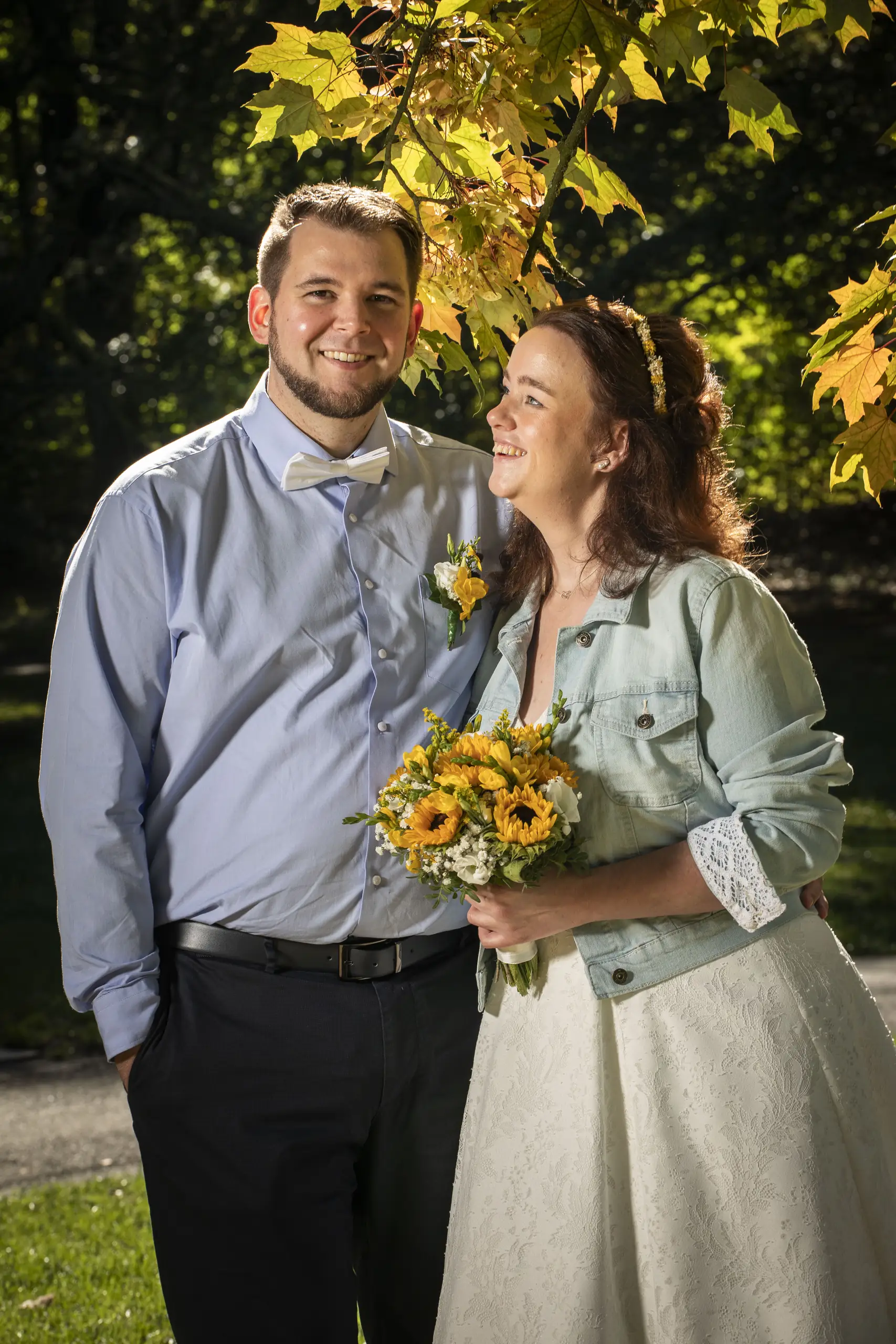 Sandra und Norman mit Sonnenblumenstrauß im warmen Licht unter Bäumen.