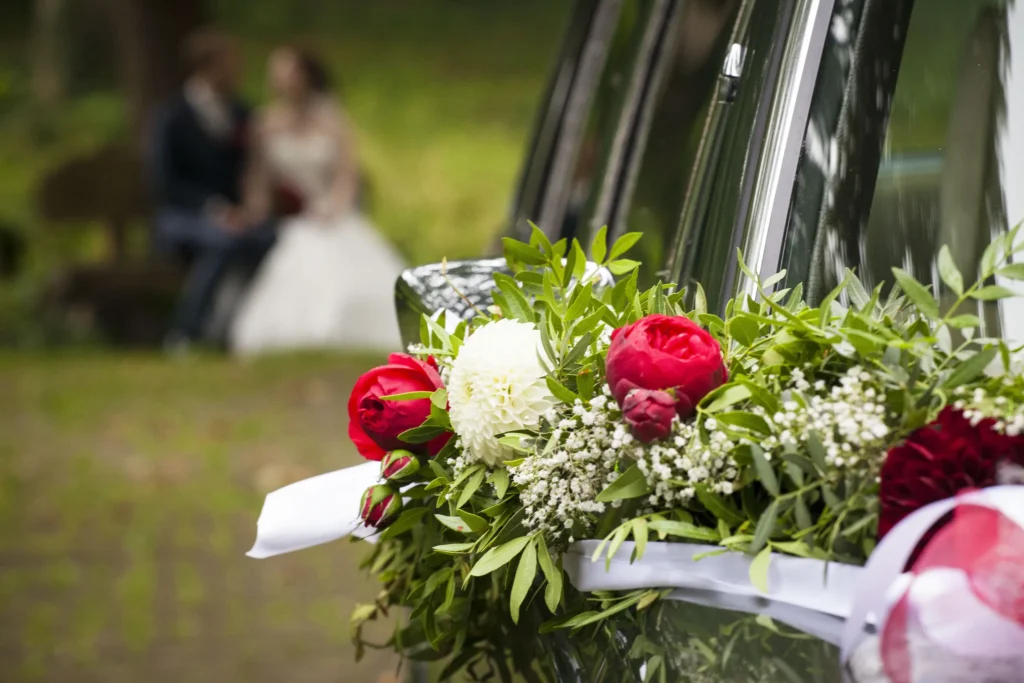 Roter und weißer Blumenschmuck am Hochzeits-Oldtimer mit Timo und Franzi im Hintergrund