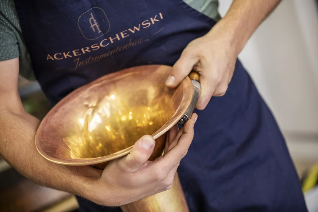Nahaufnahme von Händen, die den kupferfarbenen Schalltrichter eines Blechblasinstruments vor einer blauen Arbeitsschürze halten.