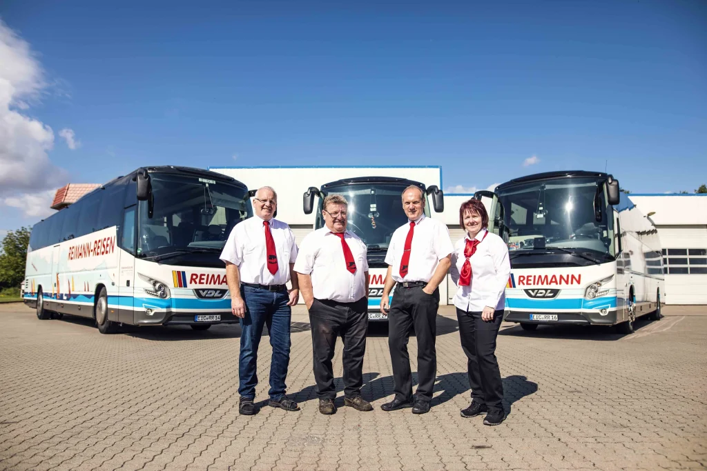 Vier Teammitglieder von Reimann Reisen in weißen Hemden und Blusen mit roten Accessoires vor drei Reisebussen.